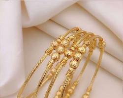 Delicate gold-toned bangles with intricate detailing and bead accents arranged on soft white fabric.